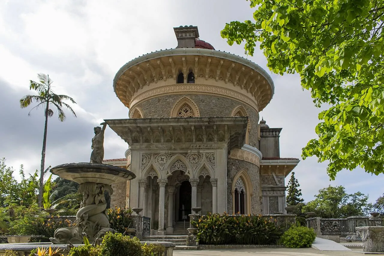 La façade du palais sous un ciel gris, son pavillon rond et son porche à arcades au-dessus d’une fontaine en pierre